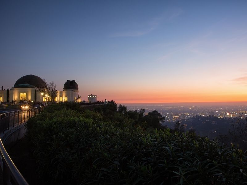 Sunset as seen from the Griffith Park observatory in Los Angeles: observatory lit up on the left side of the photo with the sunset colors and horizon in the distance.