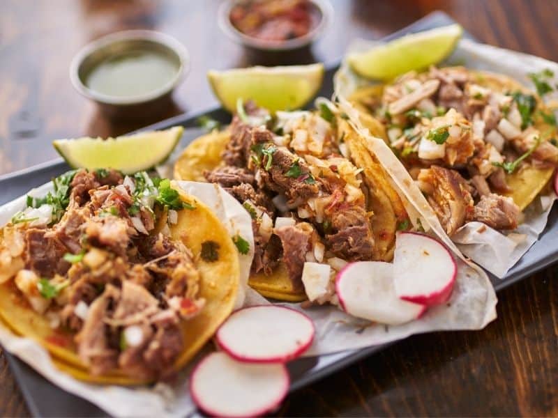 Plate of 3 tasty carnitas tacos with radish and lime and salsas on the side