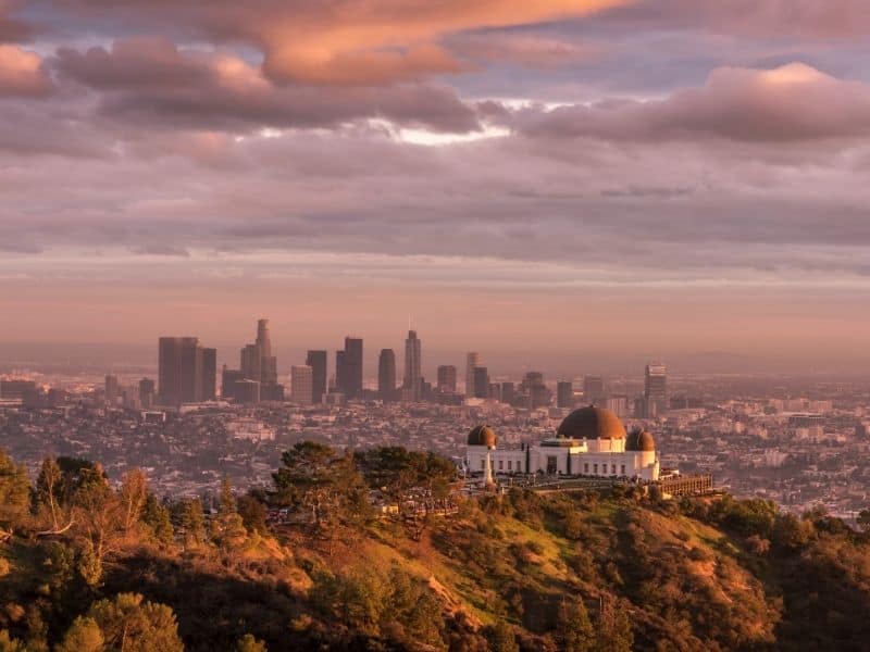 A purplish orange sky with a view of the dome-shaped Griffith Observatory in the LA hills, with the LA skyline at sunset view in the background