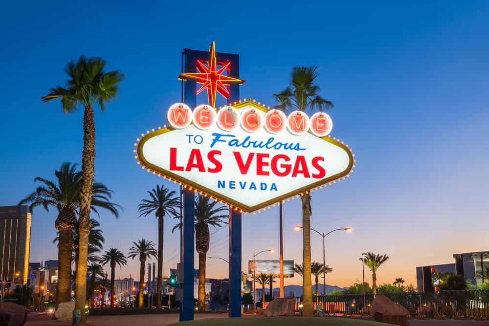 Famous nostalgic neon sign that reads "welcome to Fabulous Las Vegas Nevada" with palm trees and city lights in the background, just after sunset with orange light on the horizon and a dark blue sky.