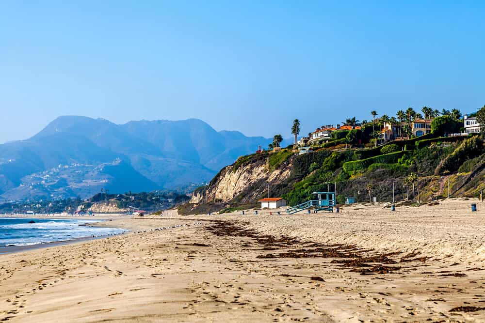 View of a seaweed-strewn beach in Southern California's beautiful Malibu, with fancy houses on the hill, a blue lifeguard box, and mountains on the coast in the background, on a sunny blue sky day.