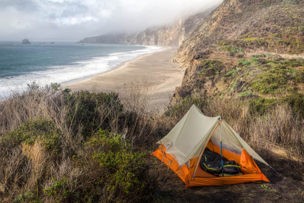 Orange tent perched on a bluff amidst lots of grass, overlooking the beautiful beach below at this Point Reyes campground near San Francisco on a slightly foggy day.