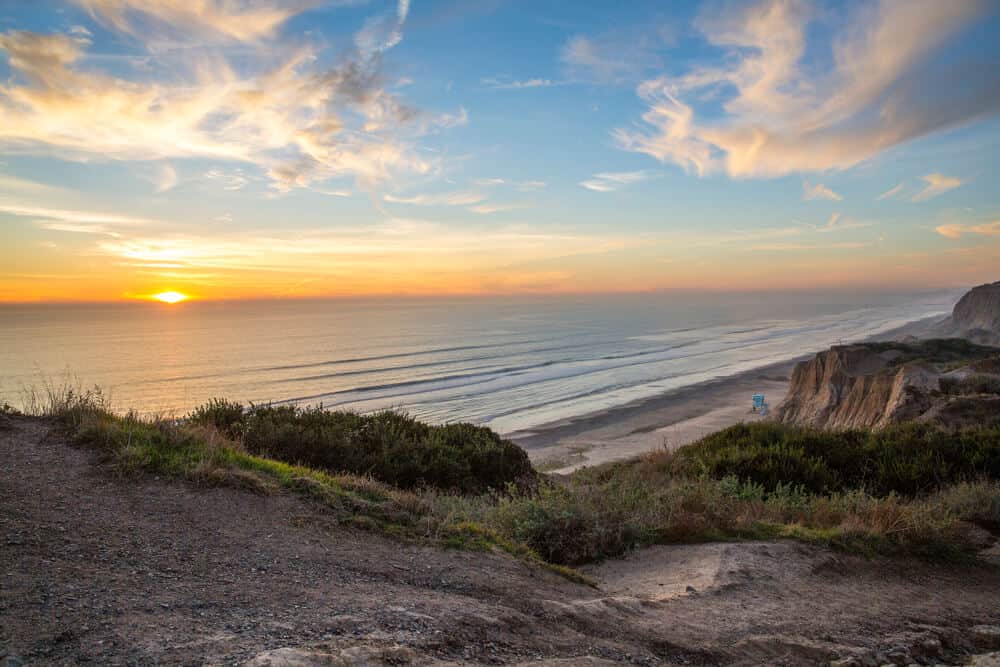 View of San Onofre from the high bluffs, cliffs leading to a beach below with one lifeguard box (baby blue) and calm water at sunset