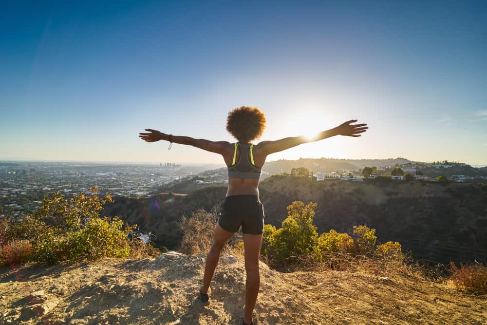 African American woman with her arms spread out, celebrating reaching the summit of this LA hike as the sun sets over the city.