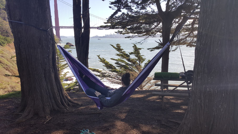 A woman reclining in a hammock with some equipment on a picnic table, overlooking the red Golden Gate Bridge and blue water.