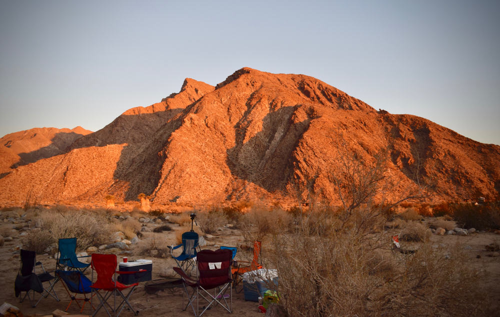 Six chairs arranged around a cooler while camping near San Diego in Anzo Borrego State Park, with the sunlight from the sunrise falling on a small hill and illuminating it orange