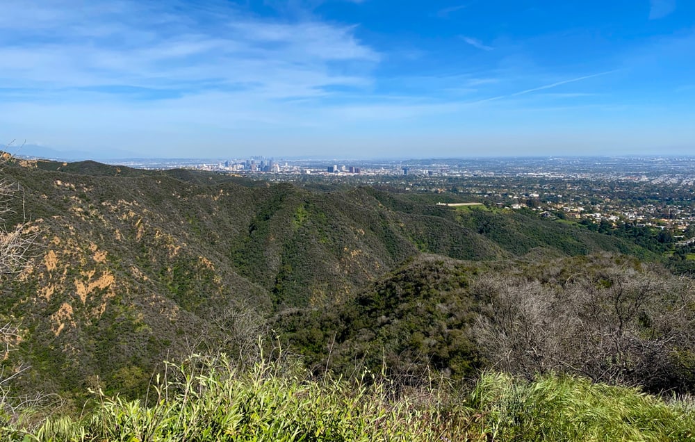 Views of Temescal Canyon, a popular LA hiking destination: green rugged hills with the cityscape small in the distant background on a sunny day.