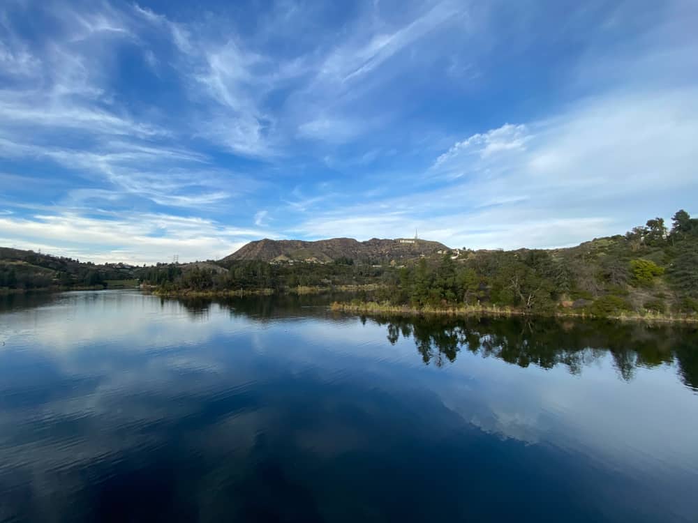 Still, glassy water in the reservoir reflecting the partly cloudy sky above, with some trees on the sides of the lake.