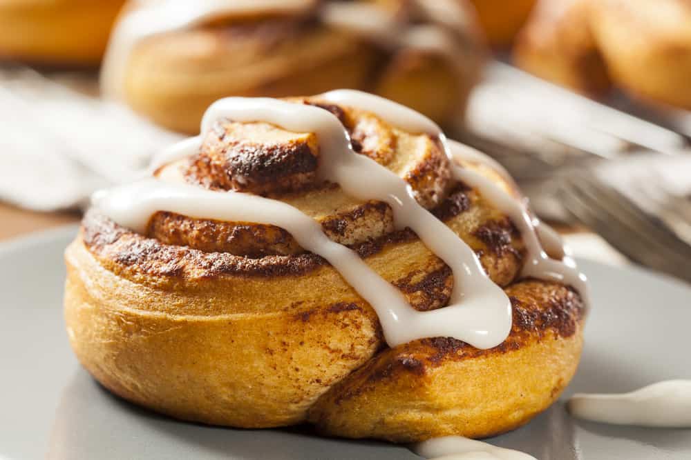 A close up photo of a cinnamon roll with white icing, others blurred in the background.