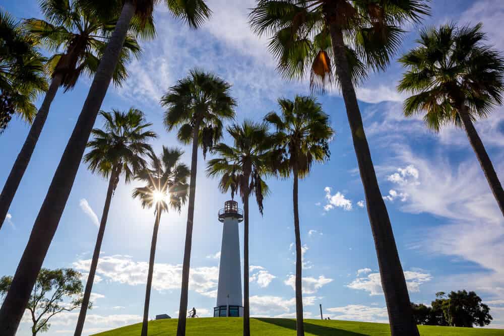 A tall white lighthouse in the center of the photo surrounded by palm trees, green grass, and a partly cloudy sky with a sunburst.