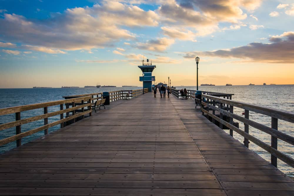 A nearly empty wooden pier at sunset with one small turquoise building and several people walking in a group in the distance, the ocean underneath the pier.