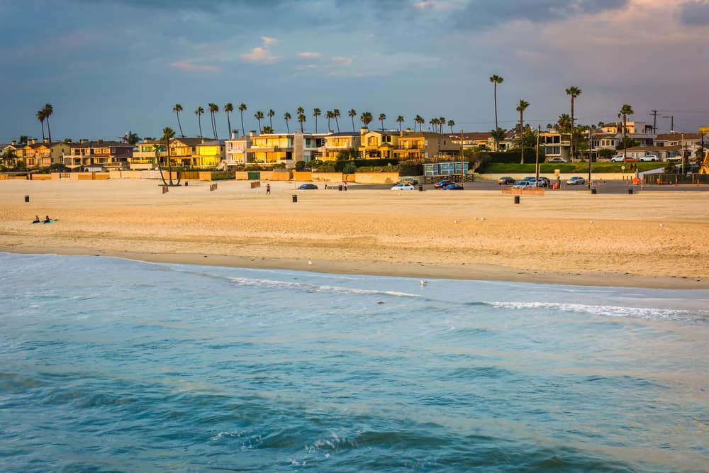 View of Seal Beach town taken from the pier at sunset, so you can see the water, sand, houses, palm trees, and colorful sky at sunset.