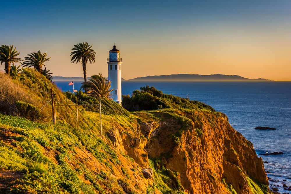 Sunset at the Point Vincente lighthouse, a white lighthouse with a red roof on a cliff in Southern California with four palm trees around it, orange horizon at sunset.