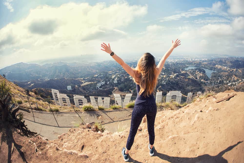 A blond woman in black athletic clothing celebrates reaching the top of the Hollywood sign, with her arms up in the air, the sign below her and the city as well.