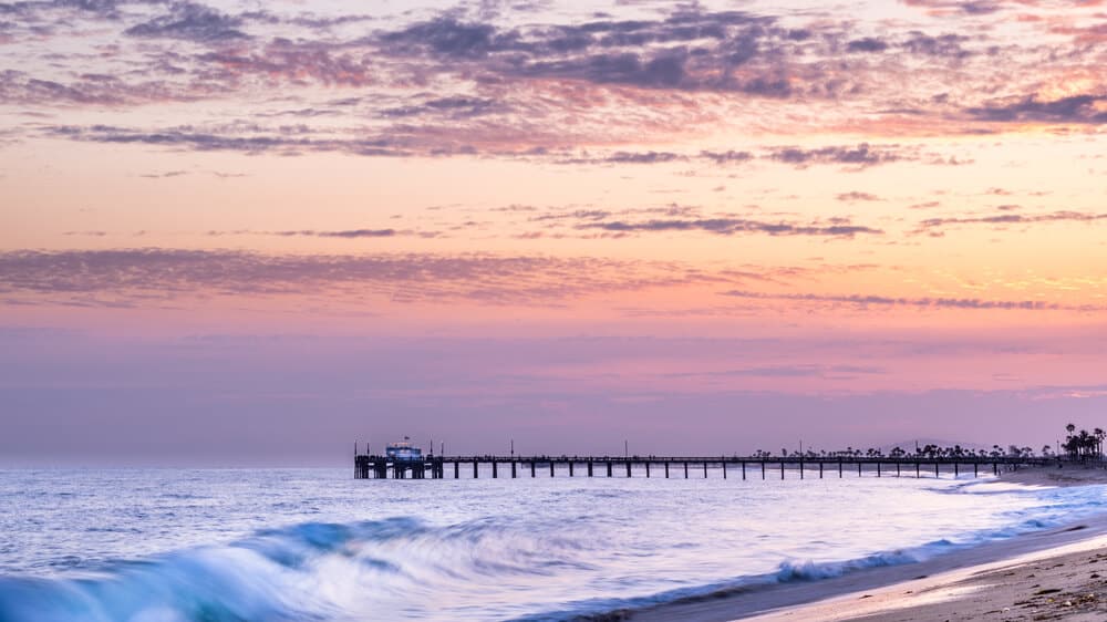 A sunset walking towards Balboa Pier, with orange, pink, and lavender tones to the sky and waves in the ocean. A sunset walk is a great thing to do in Newport Beach!