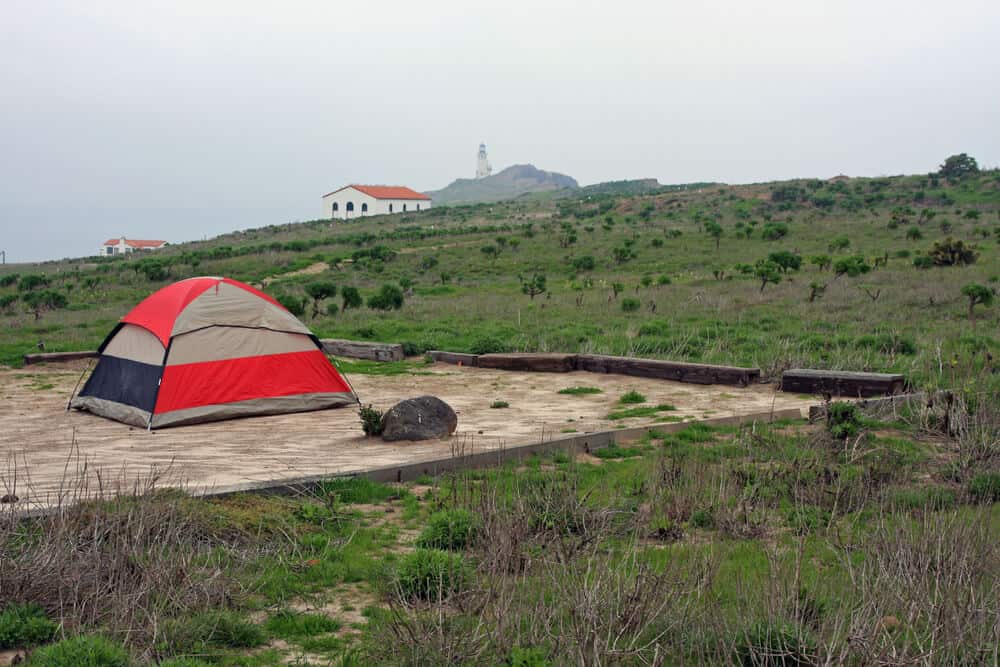 Red and gray tent in a designated campsite, surrounded by green grass and shrubs, with a lighthouse and small buildings off in the distance.