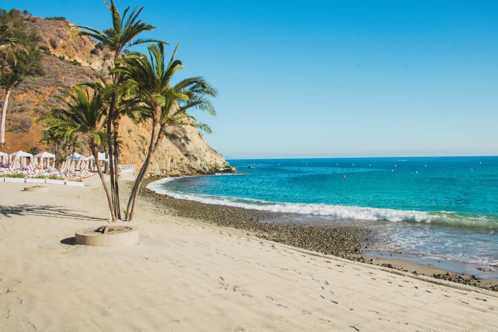 White sand beach with pebbles by the shoreline, reaching out into turquoise water, a cluster of palms and some white cabanas in the background.