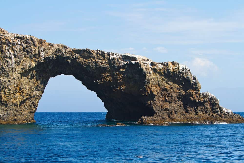 Rocky sea arch over deep blue water with a very lightly cloudy sky in the background.