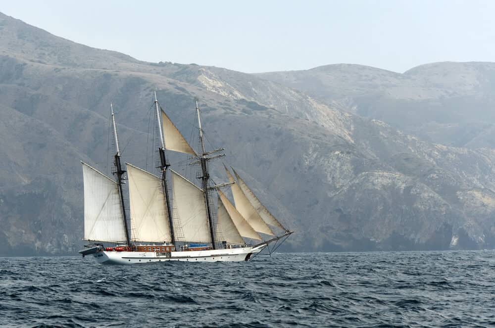 Many-masted sailboat leaving Channel Islands, in the water with island mountain in the background.