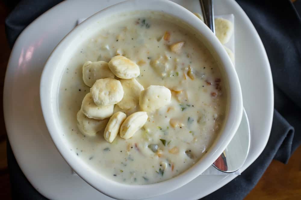 White bowl on a white plate filled with clam chowder and topped with oyster crackers with a spoon on the side.