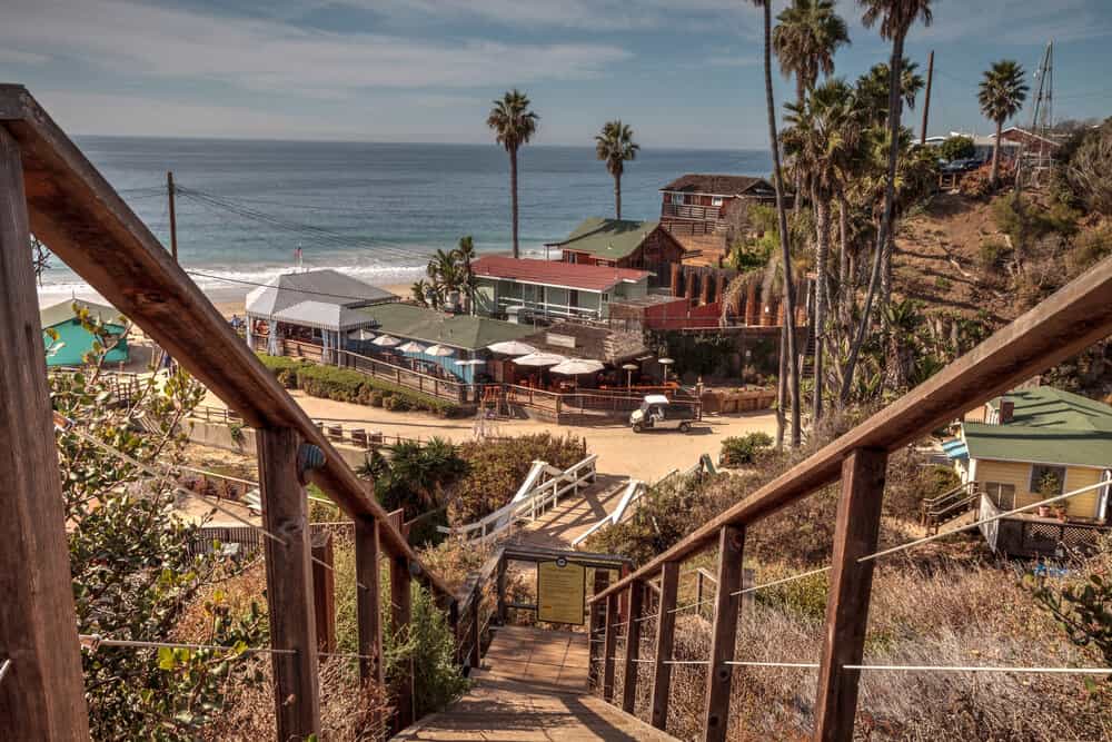 Stairs leading down to the oceanfront at Crystal Cove, surrounded by a few beachfront cottages and palm trees on the water.