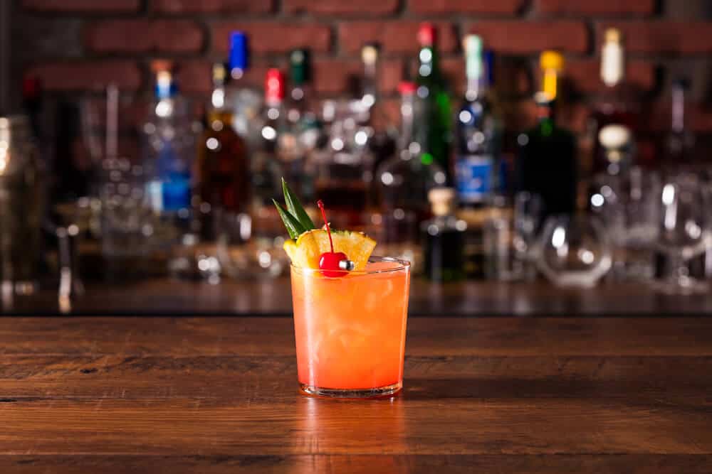 An orange and pink mai tai cocktail with a cherry and a wedge of pineapple in front of a blurry bar background.