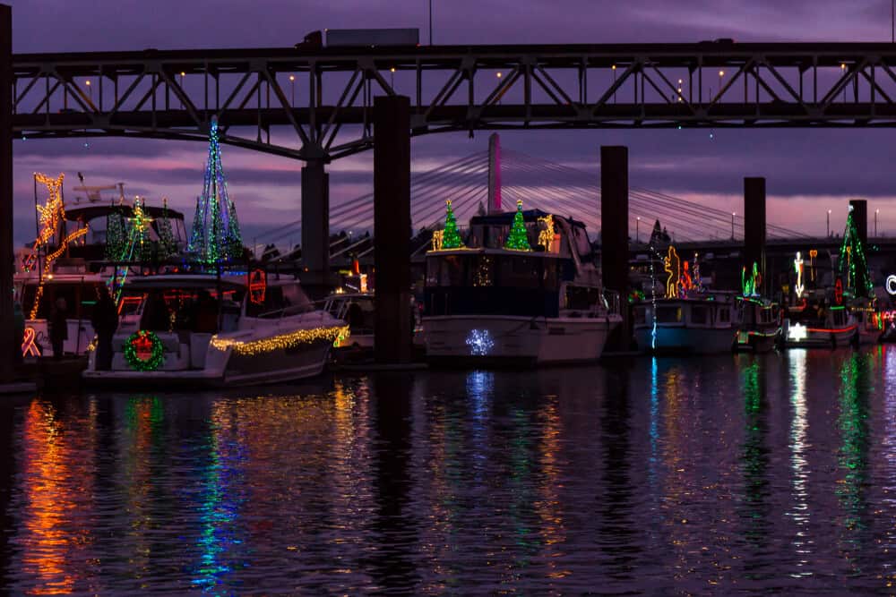 Several boats with Christmas lights on the water at sunset, with a purple and pink sky.