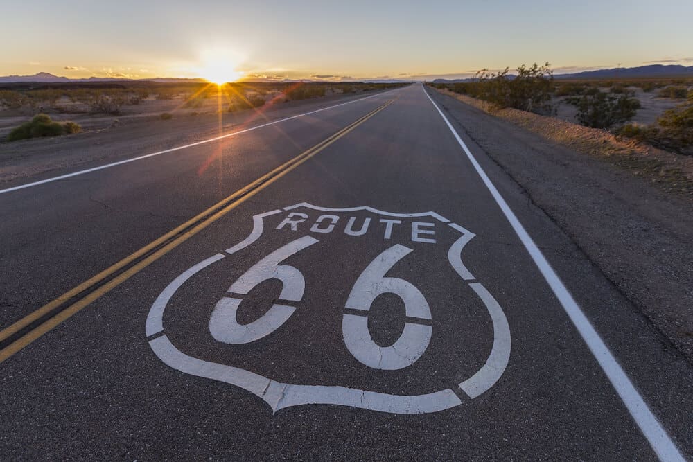 Road sign painted on the side of the highway lane which reads "Route 66" at sunset with a sunburst as the sun dips below the horizon. Road is empty.