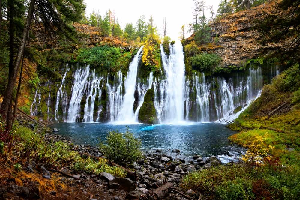 A cascading horseshoe-shape waterfall surrounded by autumn colored foliage all falling into a deep turquoise pool