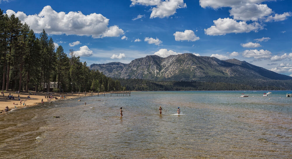 People playing in the shallow waters of Lake Tahoe in summer, other people sitting on the sandy shore of the lake, mountains and a partly cloudy sky in the background.