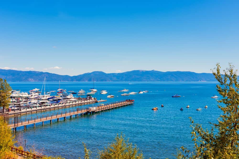 Boats docked at the Tahoe Marina and lots of smaller boats out on the lake, two wooden piers, and trees and mountains fringing the lake.