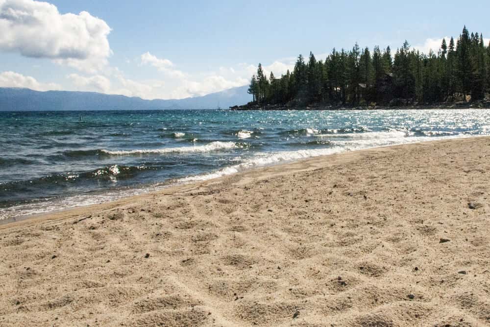 The fine sand at Meeks Bay Beach, water from the lake, and trees and mountains in the distance.