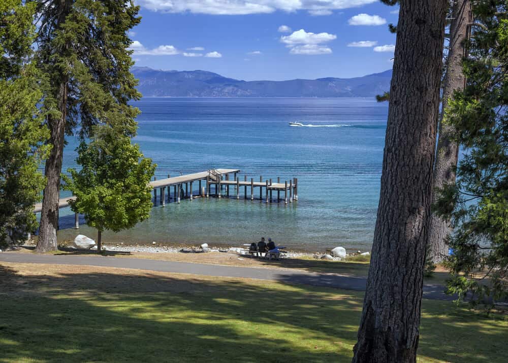 A small pier leading to a dock for boats on the shores of Lake Tahoe, a family of three sitting on a bench overlooking the lake, and mountains ringing the lake with one lone boat out on the water.