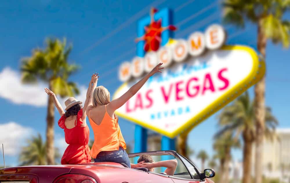 Two girls sitting atop a convertible car, after finishing a road trip to Vegas, in front of the welcome to fabulous las vegas sign.