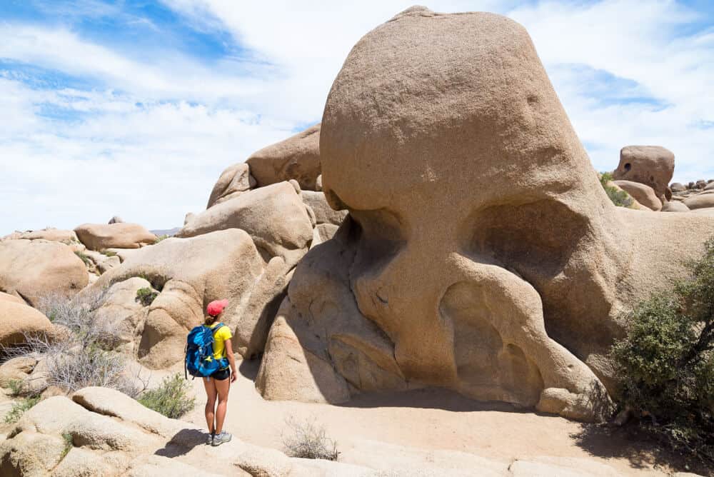 Woman in red hat and blue backpack looking at the beige rock formation that looks somewhat like a skull on a partly cloudy day in Joshua Tree.