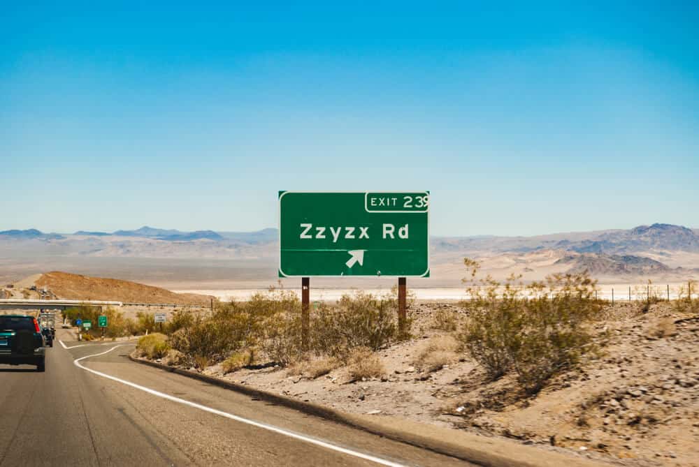 A green street sign with white letters which reads "Exit 23 Zxyxx Road" with some other cars on the highway out in the desert.