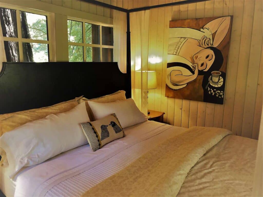 Cozy black bed with white sheets with detail of pillows and blankets on tp, white walls with a painting on it and trees outside the window