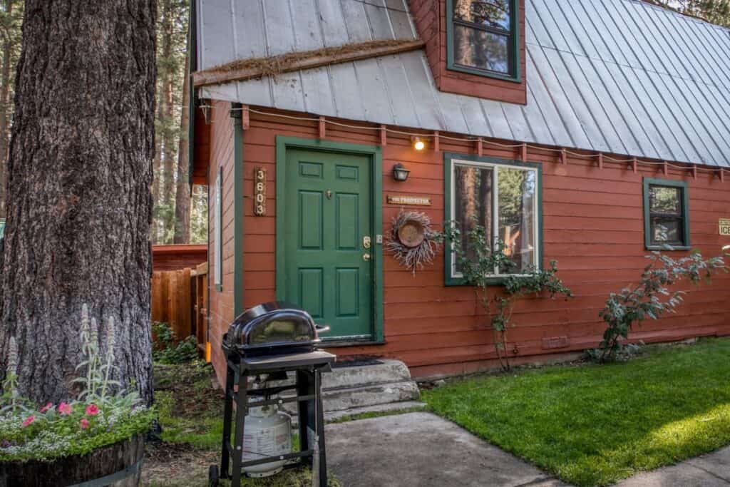 A charming pet friendly cabin in Tahoe with wood detailing, a green door, a BBQ grill in front and a small grassy area.