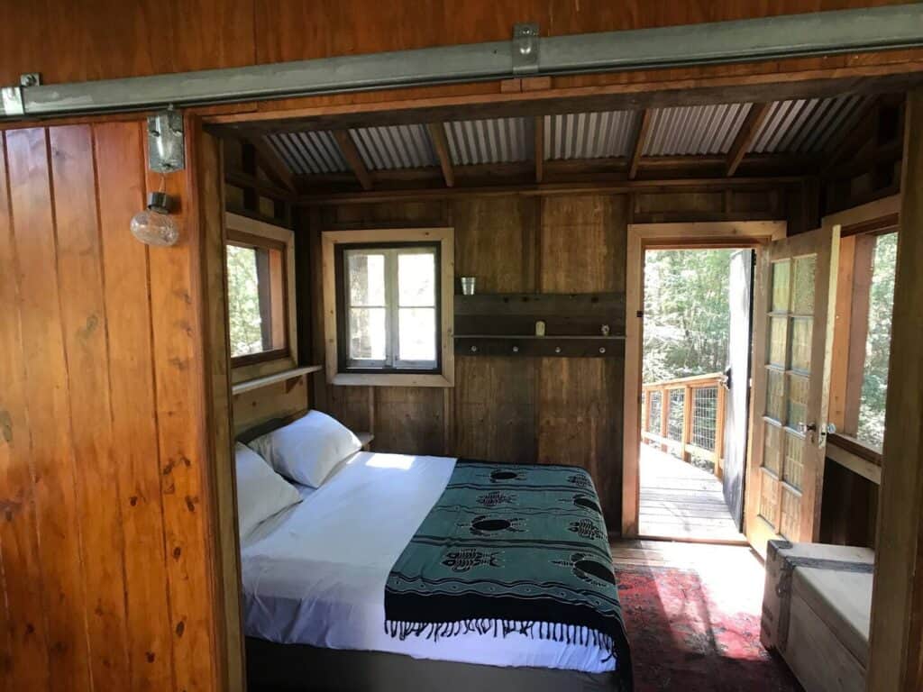 White bed with a green blanket in a wooden tree house with a metal roof with the door open showing a forest outside the treehouse.