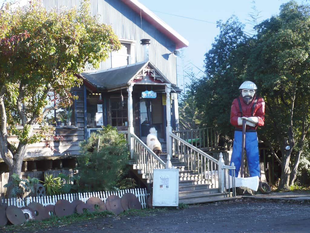 Statue of a man with a big ax wearing a red shirt and blue jeans and suspenders in front of the Blue Ox Millworks Museum. Dog on the staircase of the entrance.