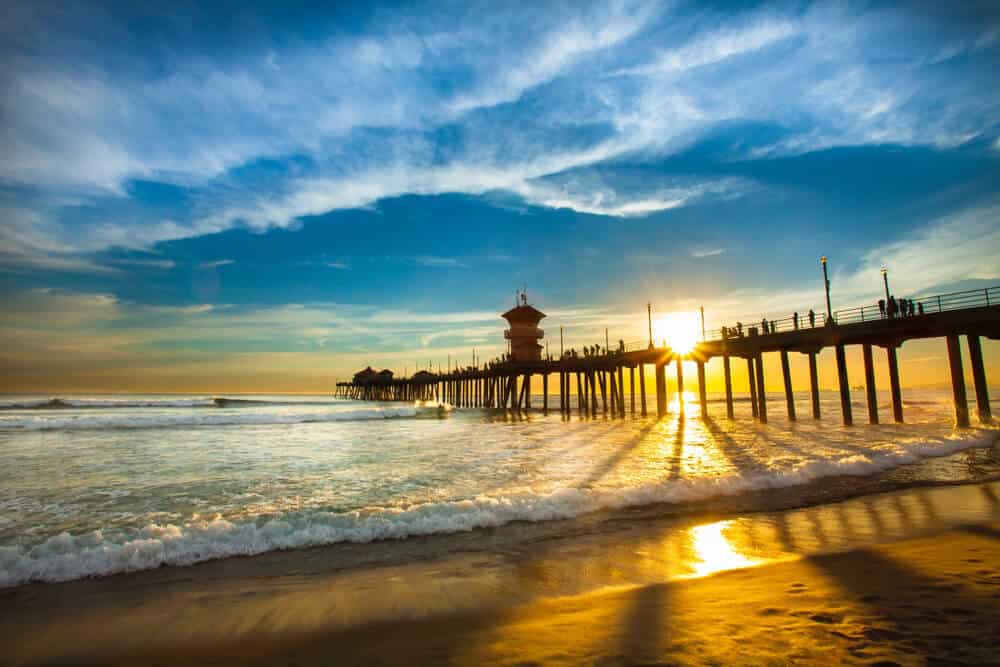 The famous Huntington beach pier at sunset with the sun sinking below the pier with a cloudy sky.