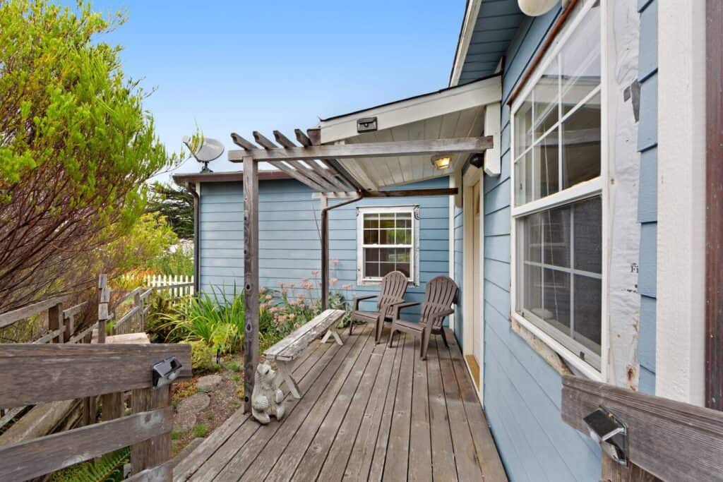 The exterior of a baby blue Mendocino airbnb cottage with a wooden porch with two chairs surrounded by plant life.