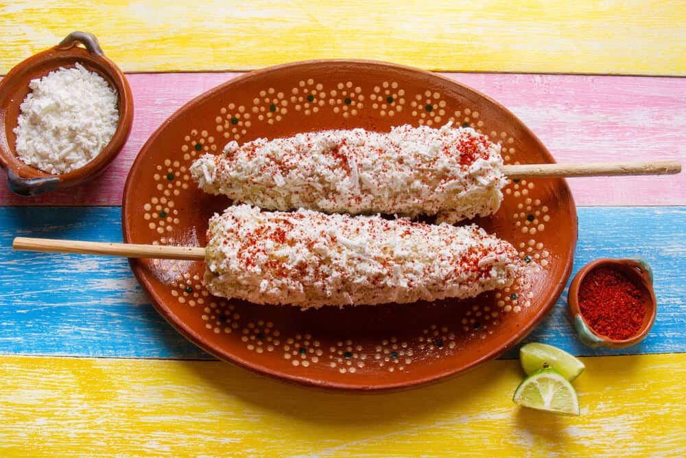 Two pieces of elote (Mexican street-style corn) on a colorful pink, yellow, and blue table.