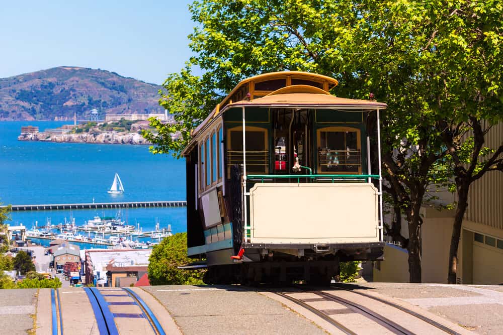 A cable car on Hyde Street going down a steep hill towards the Bay with the island of Alcatraz visible in the distance, as well as a sailboat and a marina.