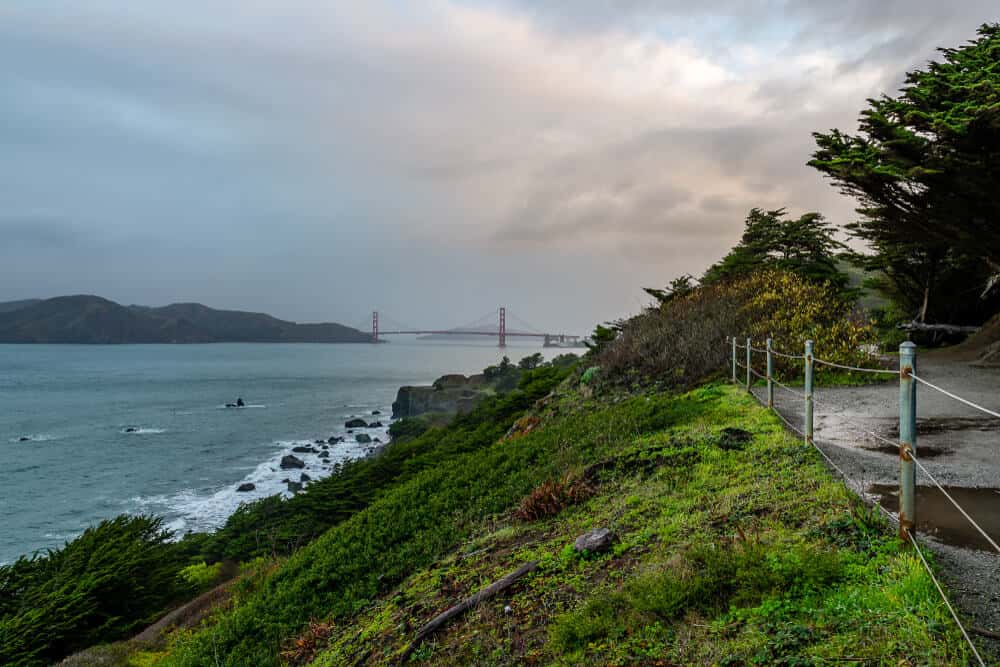 Rocky trail leading towards Lands End with a view of the red bridge of Golden Gate Bridge in the distance.