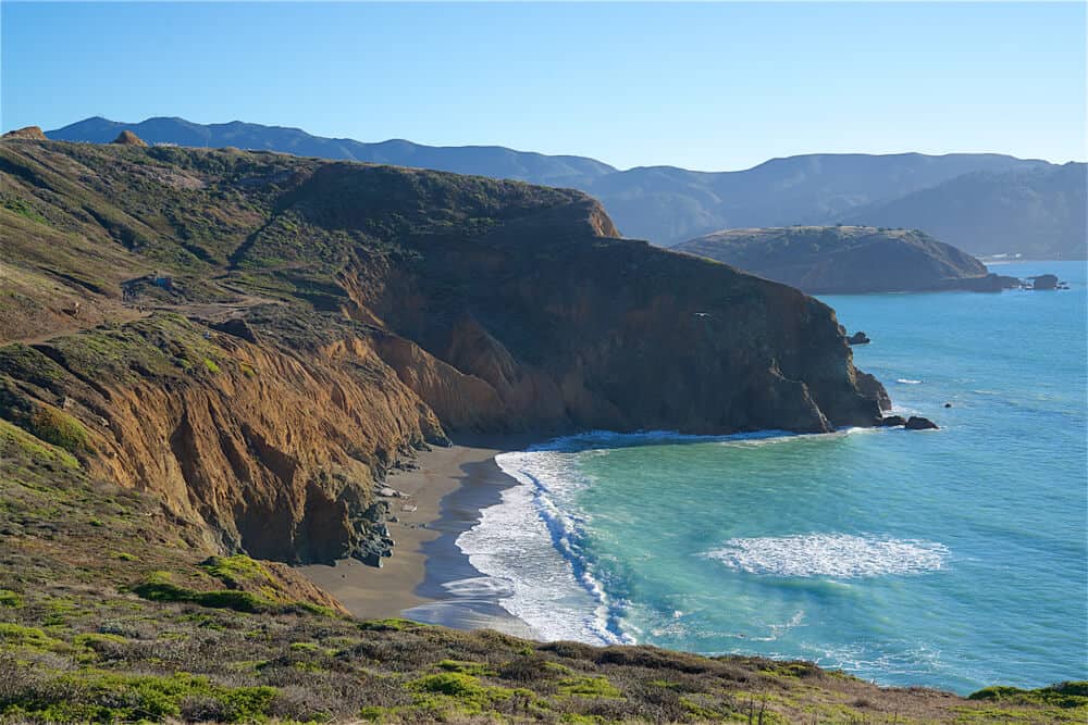 A black sand beach beneath reddish coastal bluffs, with brilliant turquoise water of the Pacific Ocean, on this beloved Bay Area hike to Mori Point.