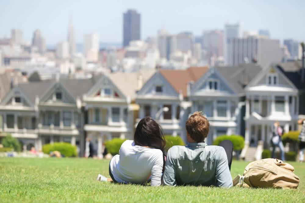 A woman and a man wearing long sleeved shirts and jeans sitting in Alamo Square overlooking the famous Painted Ladies houses and San Francisco skyline.