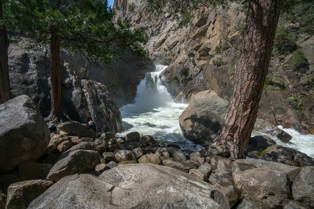 The rushing waters of Roaring River Falls Trail, a popular hike inKings Canyon, surrounded by rocks, water, and tree trunks.