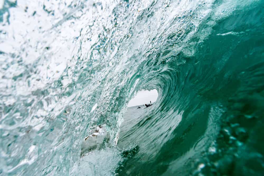 Inside the barrel of a greenish-turquoise wave, with surfers visible through the end of the barrel.