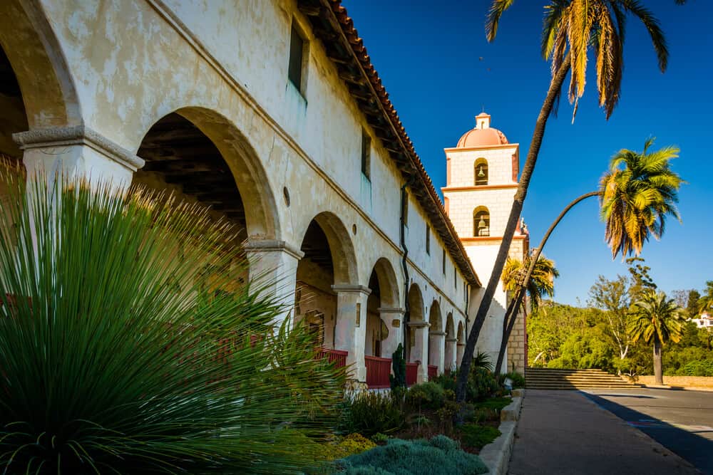 the mission at santa barbara with palm trees, plants, and spanish colonial architecture with archways and a bell tower